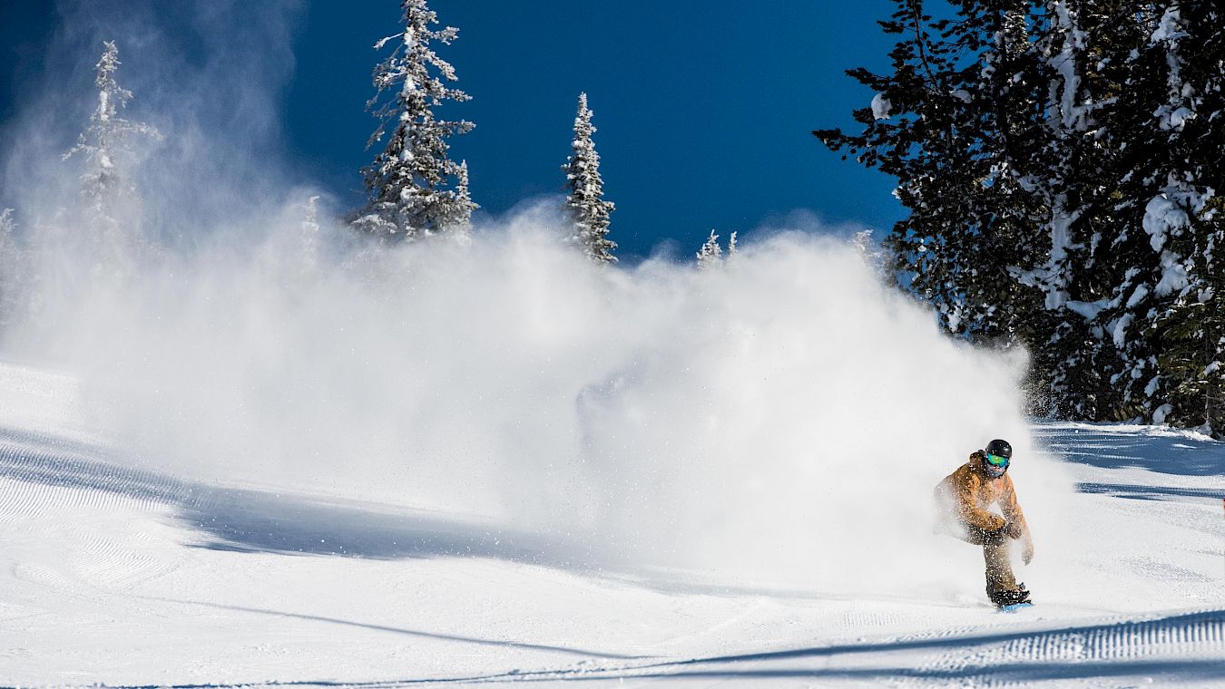 Man snowboarding down groomed run in a yellow jacket with snow cloud behind him